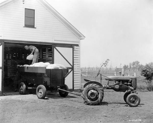 Farm manager R.M. Hawse standing in a Weber Wagon loaded with bags. The wagon is hitched to a Farmall A tractor. The bags are labeled "concrete cement" but may be filled with some other material.