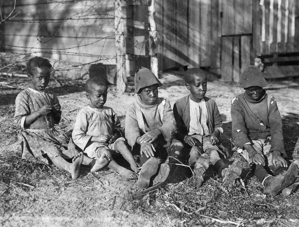 Five impoverished and disheveled-looking African American children sitting on the ground near their home. Original caption reads: "These five little Negro children show the conditions under which most southern children are brought up. When asked what they were eating they replied 'ground peas' (peanuts). They are not near enough to a Negro school to have the chance of an education so are being raised in ignorance and abject poverty."