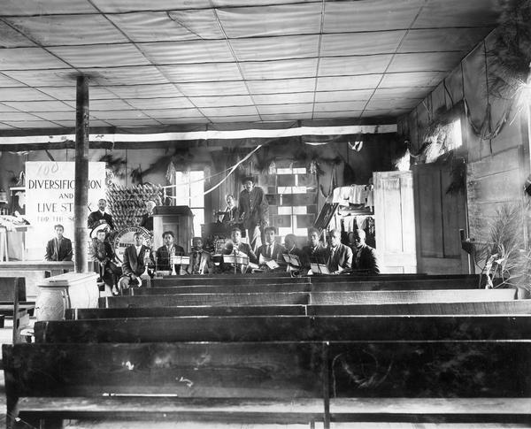 African-American school band assembled in a lecture hall at the Piney Woods Country Life School with Agricultural Extension Department workers and charts in background. Original caption notes that Professor Holden, an International Harvester Agricultural Extension lecturer, spoke at the school on March 8, 1914.
