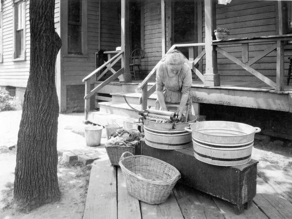 Mrs. Eshe demonstrating the washing of laundry with a washboard, ringer and tub in front of a farm house. The original caption reads: "Washing with no improved equipment. Mrs. Eshe posing."