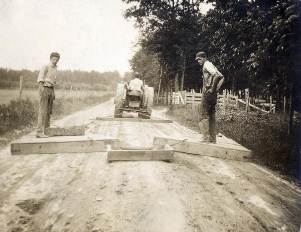 Road workers grading a rural country road with an Avery tractor and wooden grading blocks. The image was cropped from a postcard collected by International Harvester's Agricultural Extension Department to illustrate the condition of rural roads.