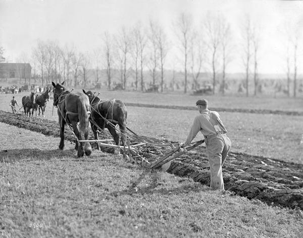 Two men appear to be competing in a plowing competition with horse-drawn walking plows. Onlookers and automobiles are in the background. The photograph was taken for, or compiled by International Harvester's Agricultural Extension Department.