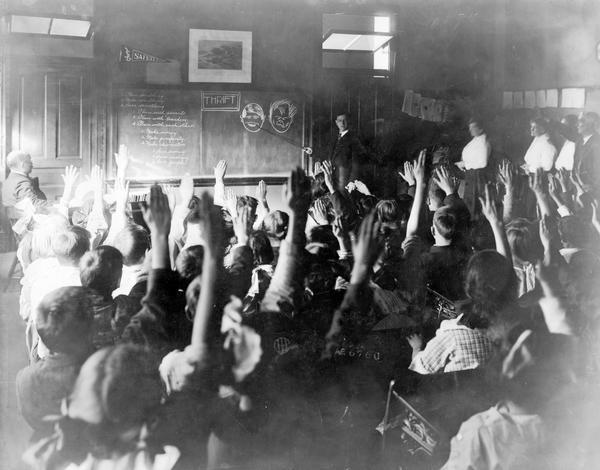 Students are raising their arms as an instructor is discussing the merits of thrift. The instructor is pointing to chalkboard illustrations of a well groomed face and a disheveled face. The room is crowded with children and teachers. Writing on blackboard reads: "Plan something. Make something. Save Something. Plan with parents. Plan with teachers. Plan with each other. Make money. Make prosperity. Make character. Save money. Save energy. Save people."