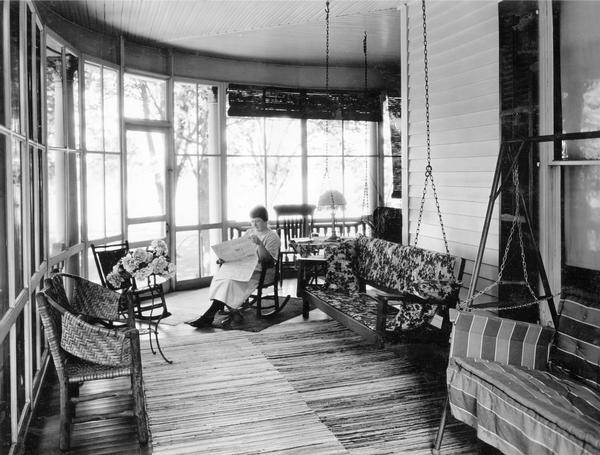 Miss Marilla Zearing reading a newspaper on a rocking chair in the screened porch of her home. The photograph was taken for International Harvester's Agricultural Extension Department. The original caption reads: "Home Economics - comfortable, screened-in porch."