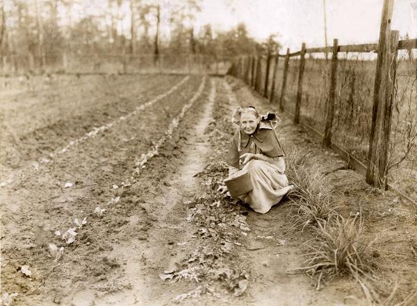 Mrs. James M. Baker in her winter garden picking turnips. Original caption reads: "She was pulling some winter turnips which behind her were multiplying. In front of her are seen two rows of new cabbage plants. Mrs. Baker cans enough vegetables and fruit to supply her family throughout the winter, but always has some fresh vegetables in the garden for greens."