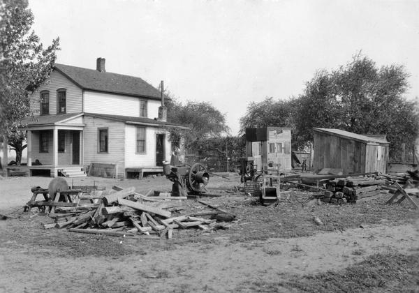 Farm house and yard "showing farmyard equipment too close to the front porch." The yard contains a stationary engine, saw, lumber, and homemade buildings.