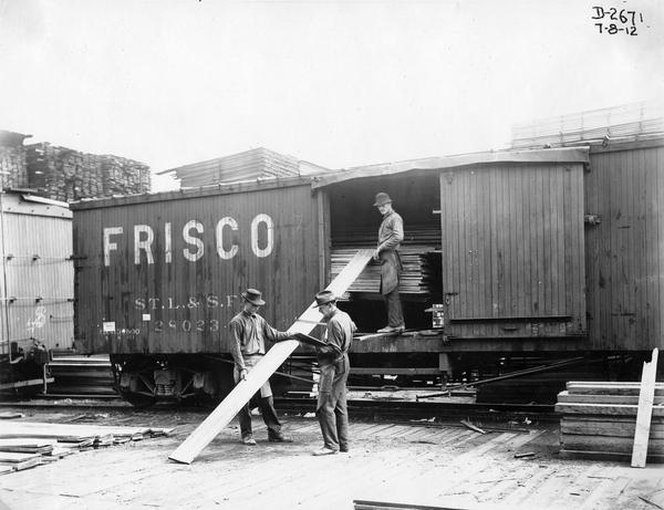 Three workers unloading planks of wood from a Frisco railroad boxcar in a lumber yard outside International Harvester's Deering Works.