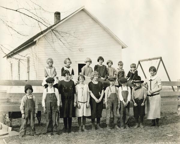 Brown Jug School students lined up along a fence outside a school house. One of the boys is wearing a cowboy hat and bandana.