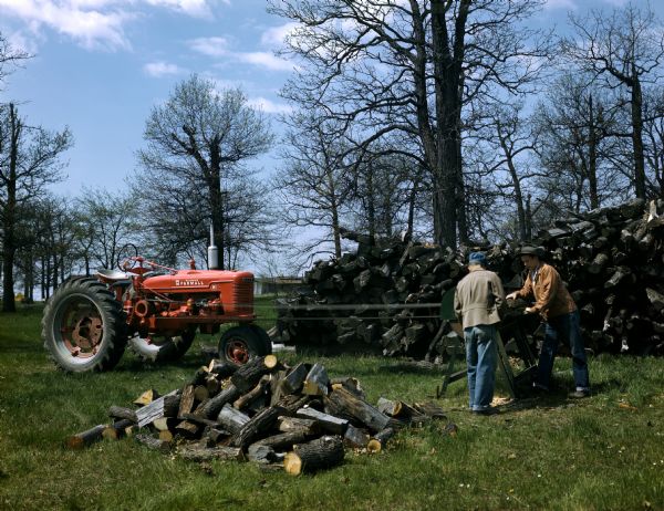 Two farmers cutting firewood with a belt-driven circular saw powered by a McCormick Farmall H tractor.