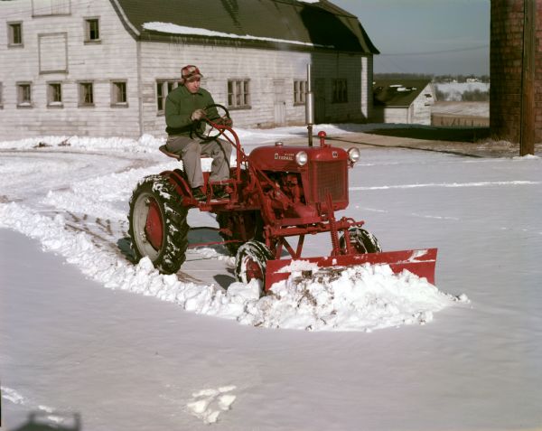 Color photograph of a farmer plowing snow with a McCormick Farmall Cub tractor equipped with a front-mounted blade.