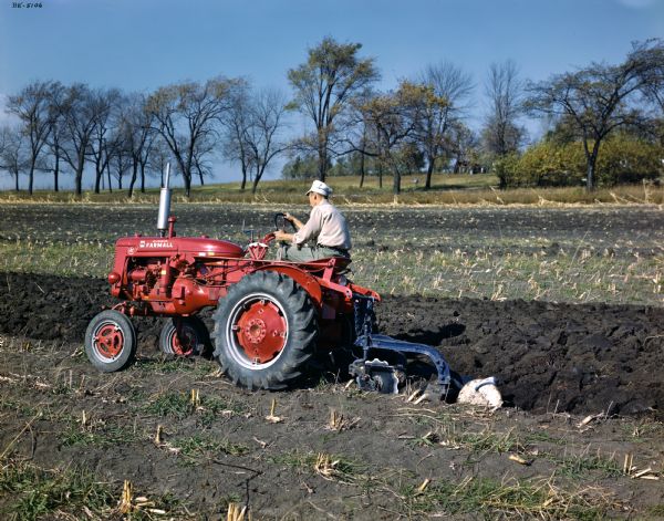 View towards a farmer plowing a field with a McCormick Farmall Super A tractor and mounted plow.