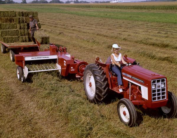 Color advertising photograph of a young woman on an International 454 tractor pulling an International 420 baler and wagon. A man is standing on the wagon.