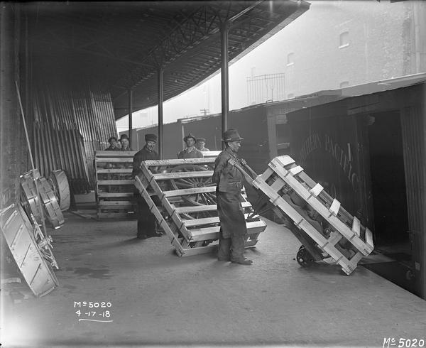 Workers loading wooden crates containing components for wagons or possibly "machine gun carts" onto railroad cars at International Harvester's McCormick Works. The crates are labeled "From Army Inspector of Ordnance; International Harvester Co.; McCormick Works." The pieces were likely manufactured under a military  contract with the U.S. government.