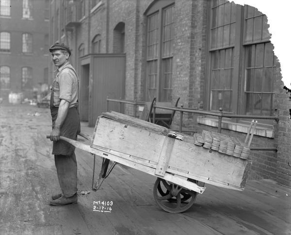 Man standing holding up the front of a wooden cart loaded with metal parts outside International Harvester's McCormick Works. The McCormick Works was built by Cyrus McCormick in 1873 and became part of International Harvester in 1902. The factory was located at Blue Island and Western Avenues in the Chicago subdivision called "Canalport." It was closed in 1961.