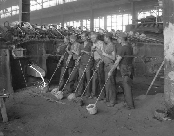 Six foundry workers lined up with pouring devices (crucibles with long handles) used for transporting molten metal at International Harvester's Auburn Works (formerly known as Osborne Works).