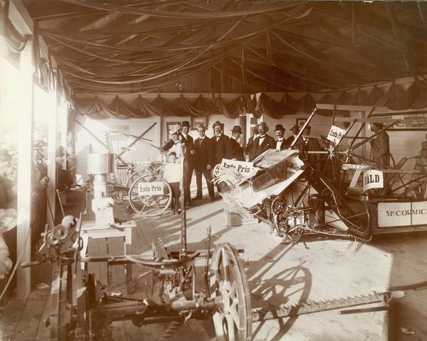 Employees of a McCormick dealership and family members standing in a pavilion with a display of McCormick farm machinery. A crowd is pressed up against the front of the display, roped off on the left, to get a better look. Two mowers, a cream separator, a grain binder, and other machines are labeled with signs reading "1st prize." The exhibition was sponsored by the McCormick dealership of Olsson and Larsson in Gefle, Sweden.
