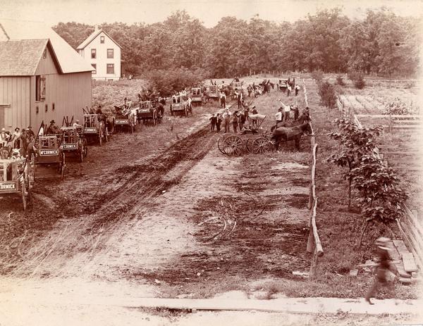 Elevated view of farmers lined up in horse-drawn wagons loaded with their new McCormick farm machines. Their wagons are adorned with "McCormick" signs and men and boys look on from a dirt street.