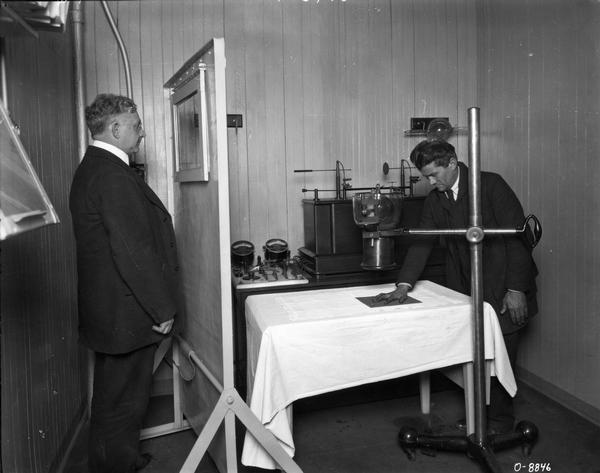 A man having his hand x-rayed as another man is looking on from behind a shield. The x-ray machine is most likely part of the medical facilities at one of International Harvester's factories (works).