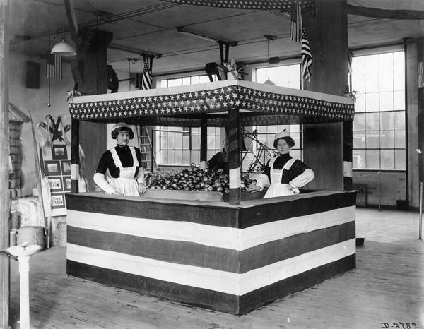 Two women staffing an apple stand for an agricultural exhibit at International Harvester's Deering Works. The stand is decorated in stars and stripes. The factory was owned by the Deering Harvester Company before 1902.