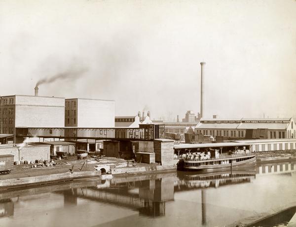 View across water towards a boat called "The Imperial" moored along the dock at the McCormick Reaper Works. The boat is loaded with freight. In the background are the factory grounds, including buildings, wagons, and rail cars. The factory was owned by the McCormick Harvesting Machine company before 1902. In 1902 it became the McCormick Works of the International Harvester Company. The factory was located at Blue Island and Western Avenues in the Chicago subdivision called "Canalport." It was closed in 1961.