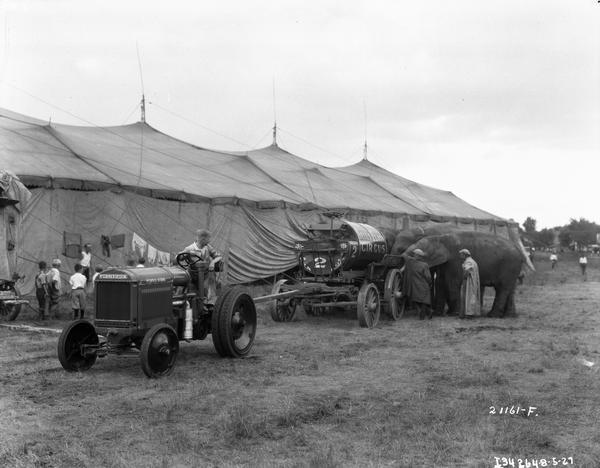 Elephants drinking water from a wagon pulled by a McCormick-Deering industrial tractor outside a circus tent belonging to the Sells-Floto Circus. Circus workers and children are standing near the tent.