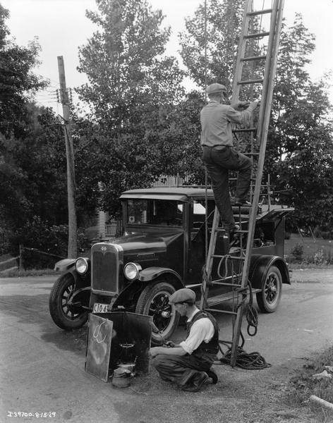 Telephone line repairmen preparing to work on a residential line. One is climbing a ladder and the other is setting up a soldering unit. Their International service truck is parked in the background.