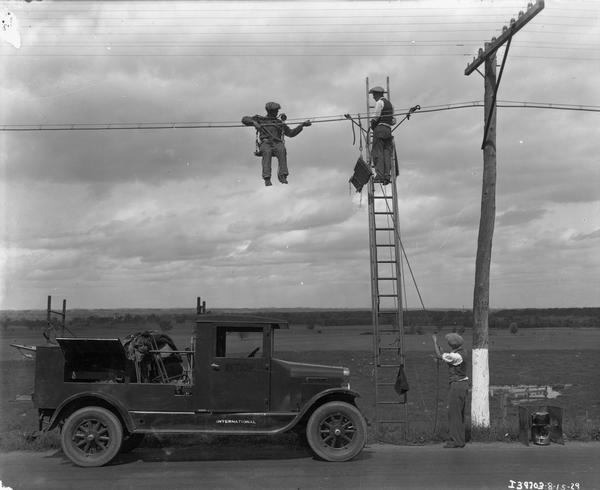 A crew of telephone line repairmen working on a rural line with their International truck parked below them. An IH truck, with storage compartments sitting open, is parked on the side of the road. On the vehicle the company name "B.T. Co." and the "International" logo is visible. A man in a harness and pulley system is hanging above the truck while another man stands on a tall ladder. A third man on the ground is leveraging ropes to stabilize the ladder. There is a lamp sitting on the ground to the right of the men. The men may be hanging street lamps.