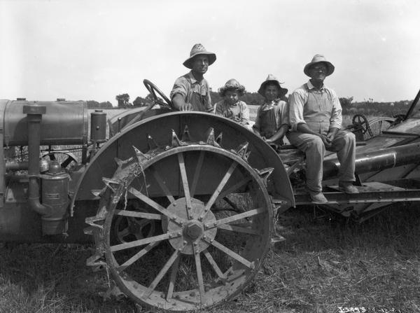 Two men and two young boys wearing overalls and straw hats are sitting on a Farmall tractor and combine (harvester-thresher) in a field.