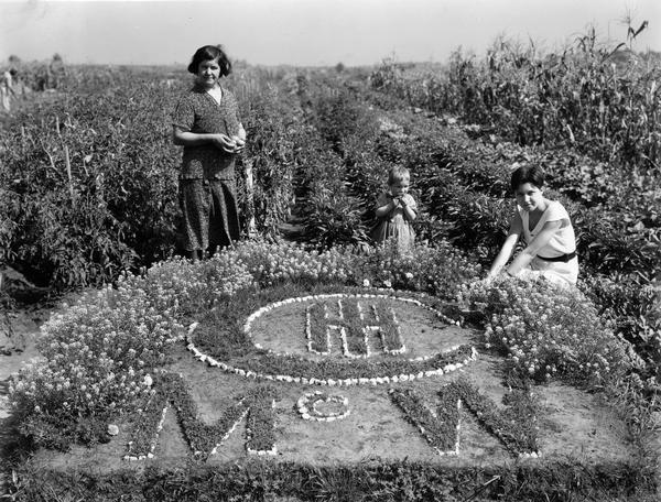 Mother and her two daughters posing near a version of International Harvester's logo at the McCormick Works community garden. The garden was one of several factory gardens created under International Harvester's "garden project." The gardens were intended to aid unemployed and underemployed workers from the company's factories. Employees with at least five years service were eligible for garden plots. The McCormick Works community gardens were located near 95th street and Crawford Avenue, and consisted of approximately 440 acres divided into 50 by 150 feet plots. The gardens were utilized by over 1,800 employees.