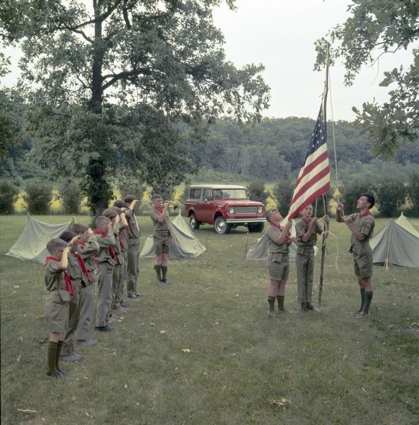 Color photograph of Boy Scouts raising a flag as others stand at attention during a ceremony. Tents and a 1972 International Scout pickup are in the background.