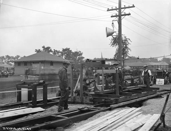 Three workers using a portable sawmill to cut wood, possibly at a fairgrounds. A McCormick-Deering industrial tractor is in the background.