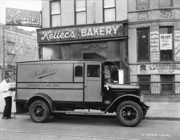 View across street towards a man in a white coat removing baked goods from the back of an International delivery truck parked in front of a storefront. The truck was owned by Keller's Bakery of Brooklyn and New York.
