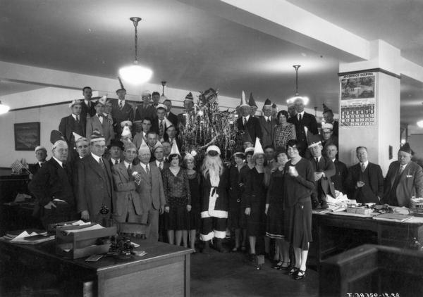 International Harvester employees standing around a Christmas tree and Santa Claus during an office party. Many of the workers are wearing party hats and some are holding American flags. The Santa appears to be wearing a mask. The party was described in "Harvester World" magazine as "the second annual Christmas party of the order and distribution department, Chicago office."