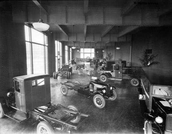 Showroom of an International Harvester factory(?) with a variety of International trucks on display. There are large show windows in the right, and in the far background.
