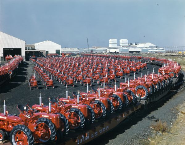 Color photograph of hundreds of Farmall tractors lined up outside the International Harvester's Louisville Works(?) (factory). Some of the tractors are loaded on railroad cars ready for shipment.