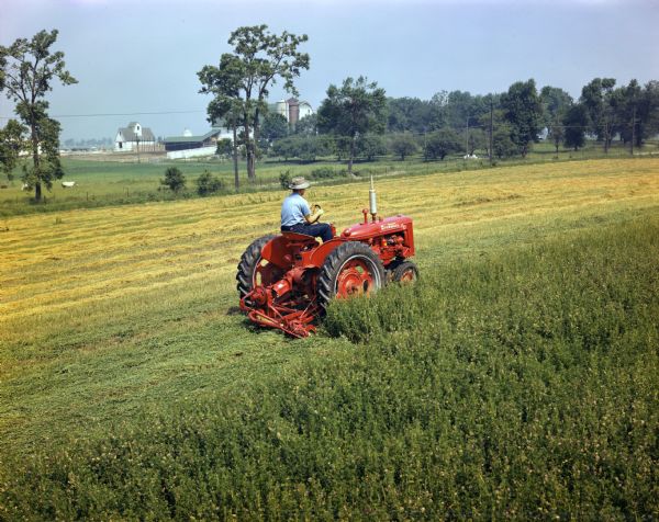 Farmall C Tractor with Mower Attachment View towards a farmer operating a McCormick Farmall C tractor with attached mower in a field. Farm buildings are in the distance beyond trees.
