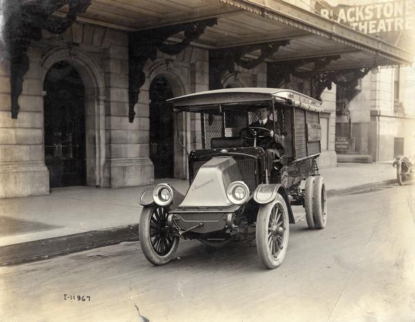 Man sitting behind the wheel of an International Model F truck parked on a city street in front of the Blackstone Theatre. The truck was owned by a Firestone agent.