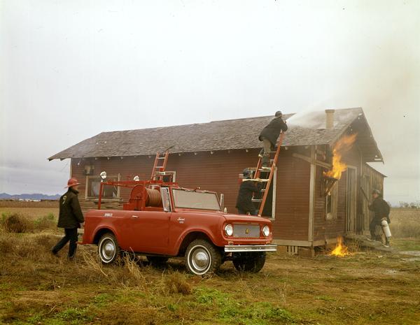 Color photograph of a fire crew battling small house fire during a training exercise. The men are using an International Scout 80 1965 fire truck.