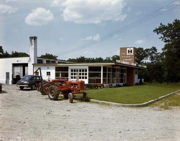 Color photograph of tractors and a car parked in front of the Fisher Company store, an IH dealership. This "prototype" dealership building was built as part of International Harvester's "Dealer Base of Operations Program." By February of 1948, 386 dealerships had been built on the prototype plan. Another 617 dealerships were under construction or had been built on a modified prototype plan. Eventually over 1800 such dealerships were built worldwide.