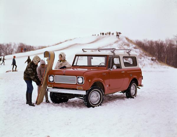 Color advertising photograph of a woman and two children with a toboggan and an International Scout 4x4 pickup truck. A snow-covered sledding hill is in the background.