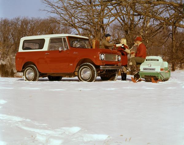 Color advertising photograph of two couples outdoors in the snow drinking cocoa(?) from a thermos next to an International Scout pickup truck and Johnson snowmobile.