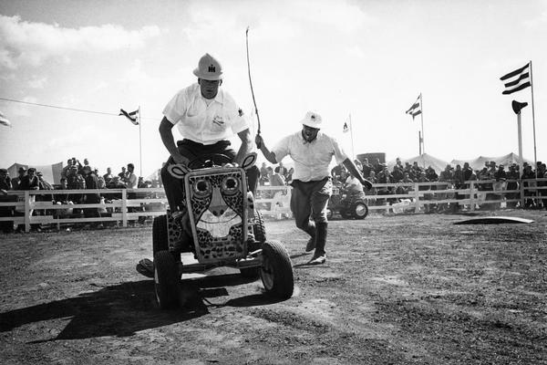 Men riding Cub Cadet lawn tractors in International Harvester's "wild-animal" act at the Farm Progress Show in Farmer City, Iowa. Three International Harvester salesmen piloted the jungle cat Cub Cadet tractors and George Mann, a professional actor, played the part of "Fisbee" the "hapless animal trainer".