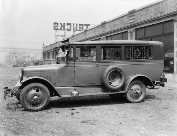 International ambulance equipped with what appears to be ornate leaded glass windows. A sign in the driver's side window reads: "United Hospital Port Chester, NY." There is also a bell on the running board. The ambulance appears to be parked near the back of an International truck dealership.