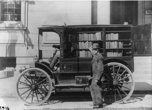 An International Auto Wagon with a rear compartment containing shelves of books is parked outside a library.  A man is sitting in the driver's seat, and a man in a bowler hat is selecting a book from what appears to be a "bookmobile."