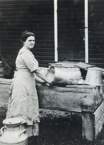 Woman standing at a table with milk cans on it. The photograph was taken for International Harvester's Agricultural Extension Department. A caption with the photograph reads: "Dairying: A Western Dairymaid; Cut 5787; E.J. Hall, Oak Park, Illinois."
