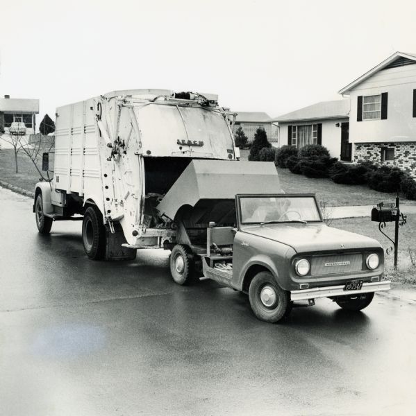 International Scout truck with dump box loading refuse into a garbage truck in a suburban neighborhood.