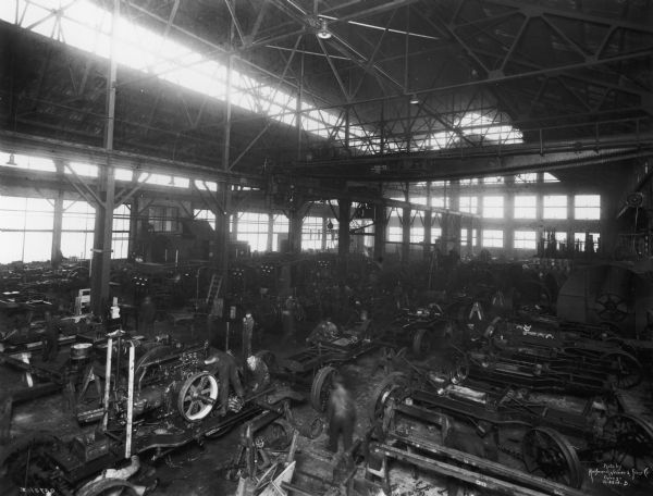 Workers on a factory floor installing engines onto tractor frames (chassis) at an International Harvester factory, most likely Tractor Works.