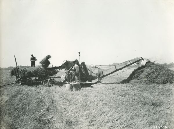 Two farmers are using a Deering harvester-thresher (combine) and horse-drawn wagon to thresh grain.