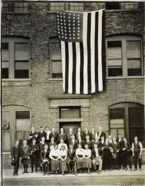 The office staff of International Harvester's Weber Works are posing beneath an American flag in front of the exterior office door. Weber Works produced wagons.