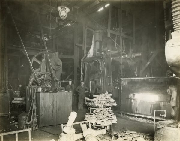 Two factory workers stand near a furnace at International Harvester's Akron Works. Piles of parts are in the foreground.